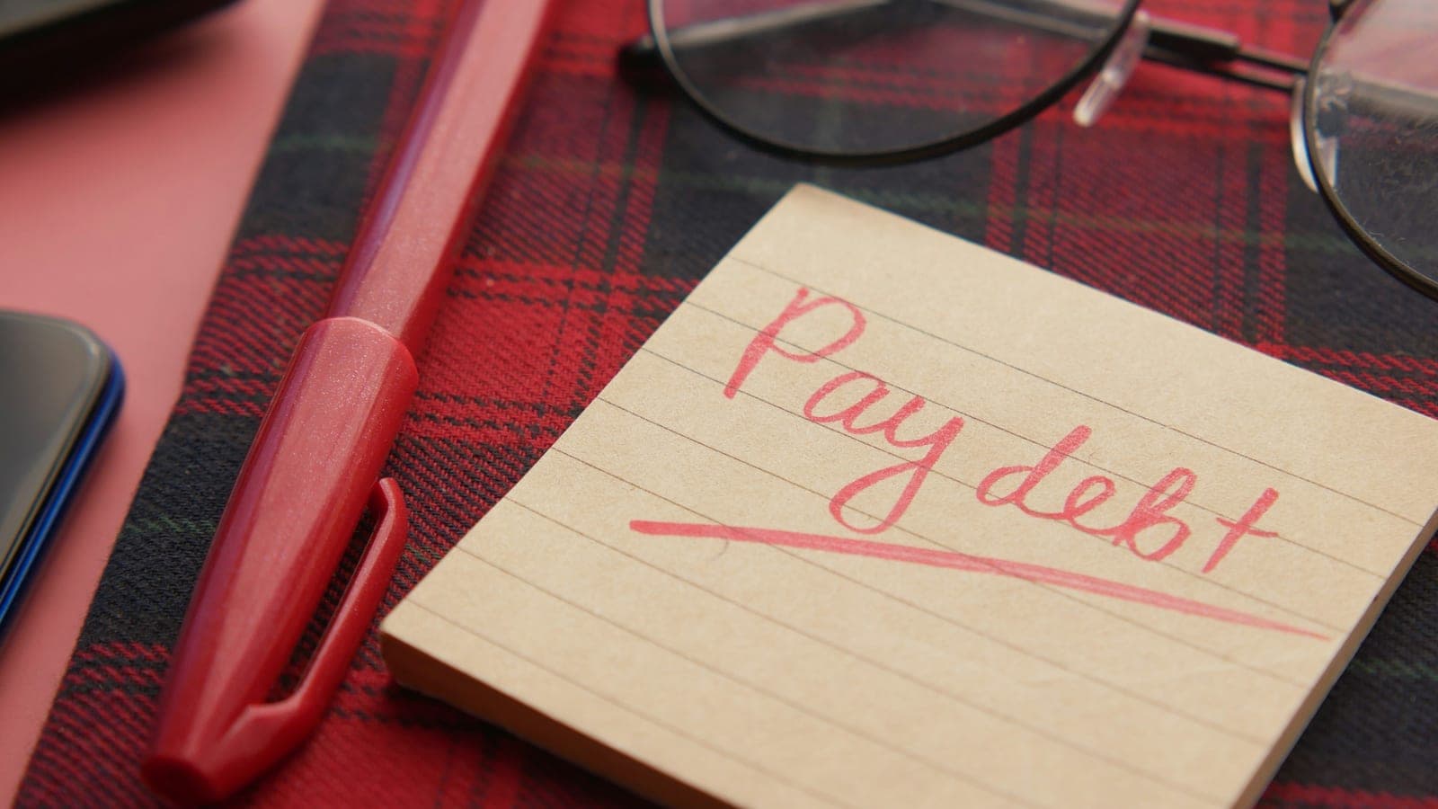 A note that says pay debt next to a pen and glasses