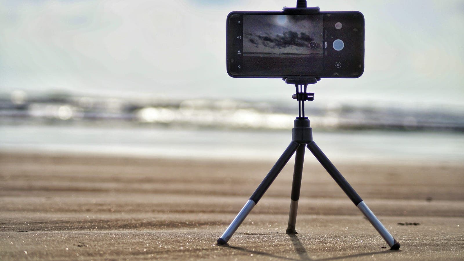 A phone mounted on a small tripod on a beach, its camera app open