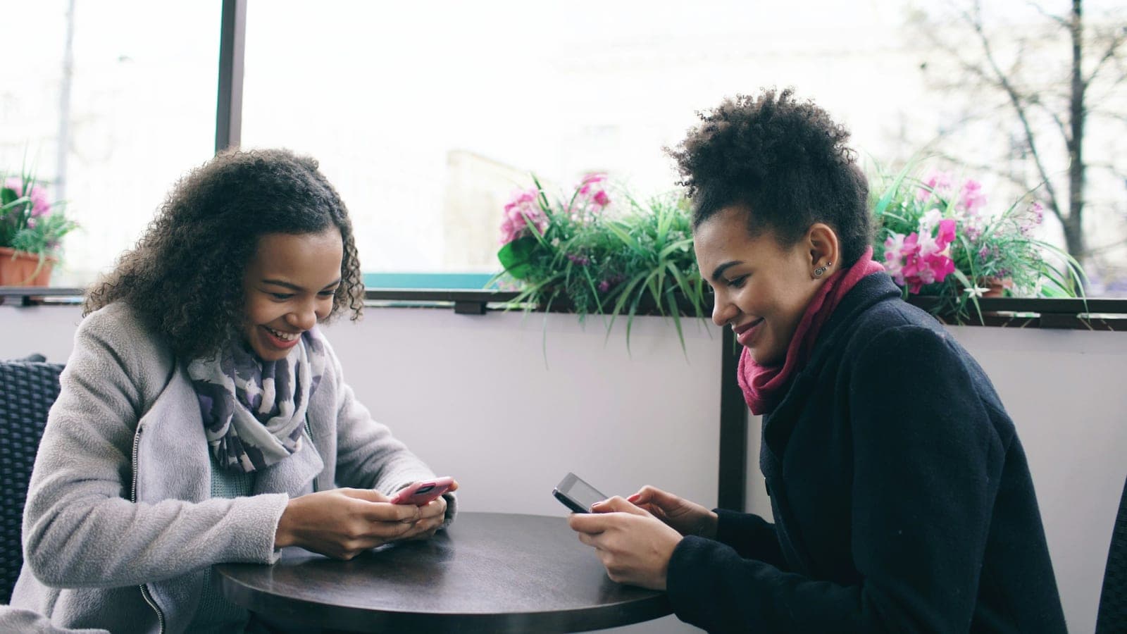 Two attractive mixed race female friends sharing together using smartphone in street cafe
