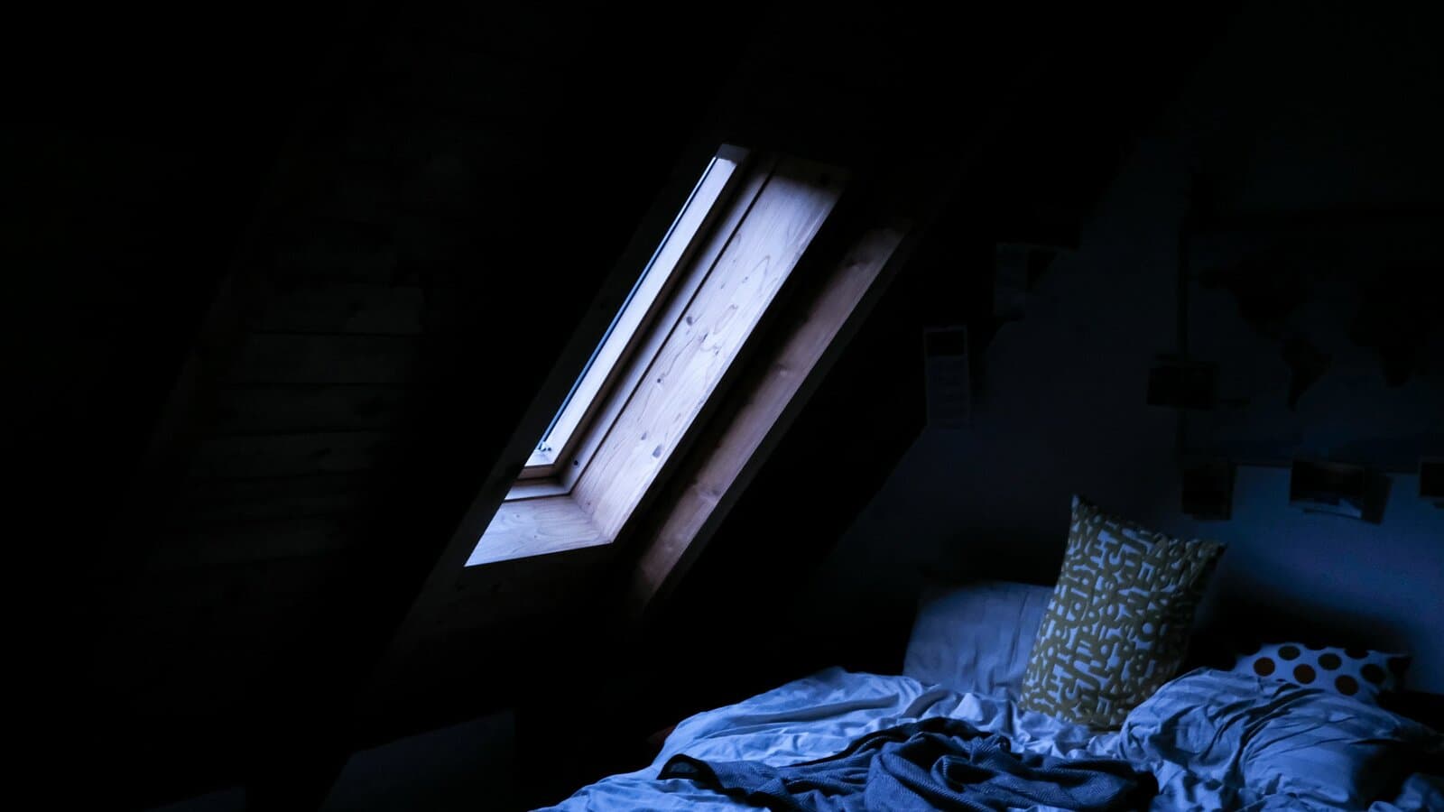 A rumpled bed under a skylight in a dark attic room