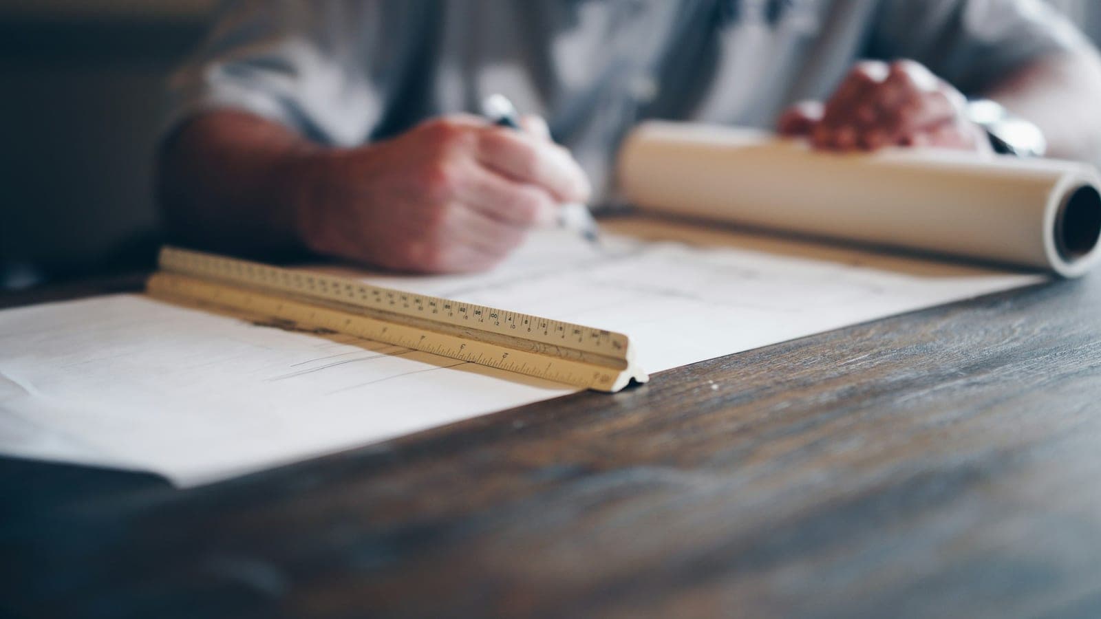 A person drafting on a technical drawing, with a scale ruler and a rolled blueprint on a wooden desk