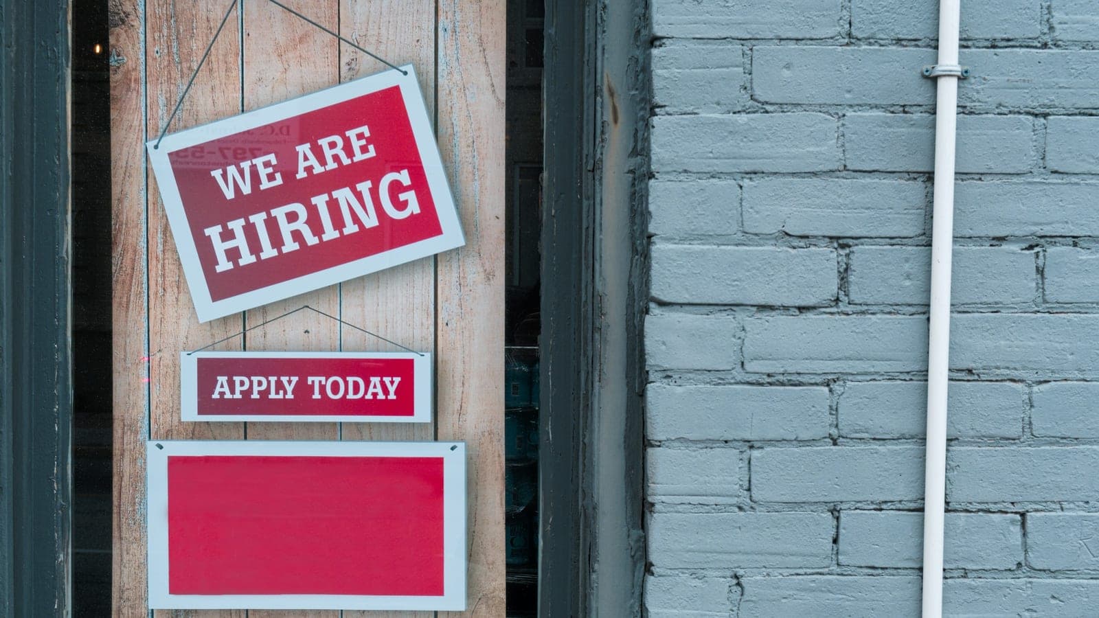 "We Are Hiring" Sign in Window of Brick Building - A red and white sign in a window of a brick building reads We Are Hiring with a smaller sign that says Apply Today.